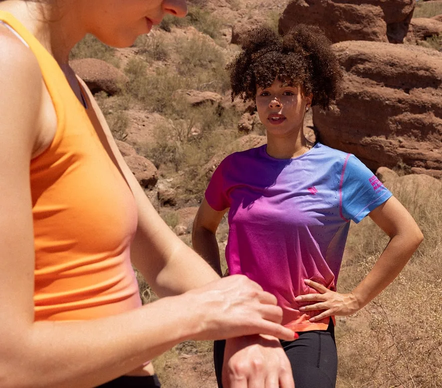 A runner wears the Every Woman's Marathon blue and pink gradient Race Pace Tee SS as she looks onward at her friend checking her watch.