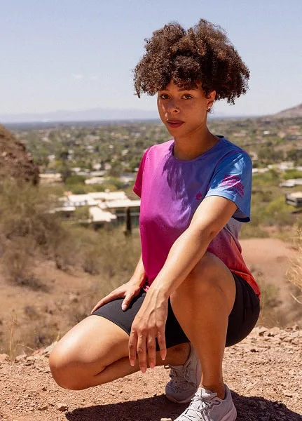 Runner wears the Every Woman's Marathon blue and pink gradient Race Pace Tee SS as she is kneeling down with the Scottsdale Landscape behind her.