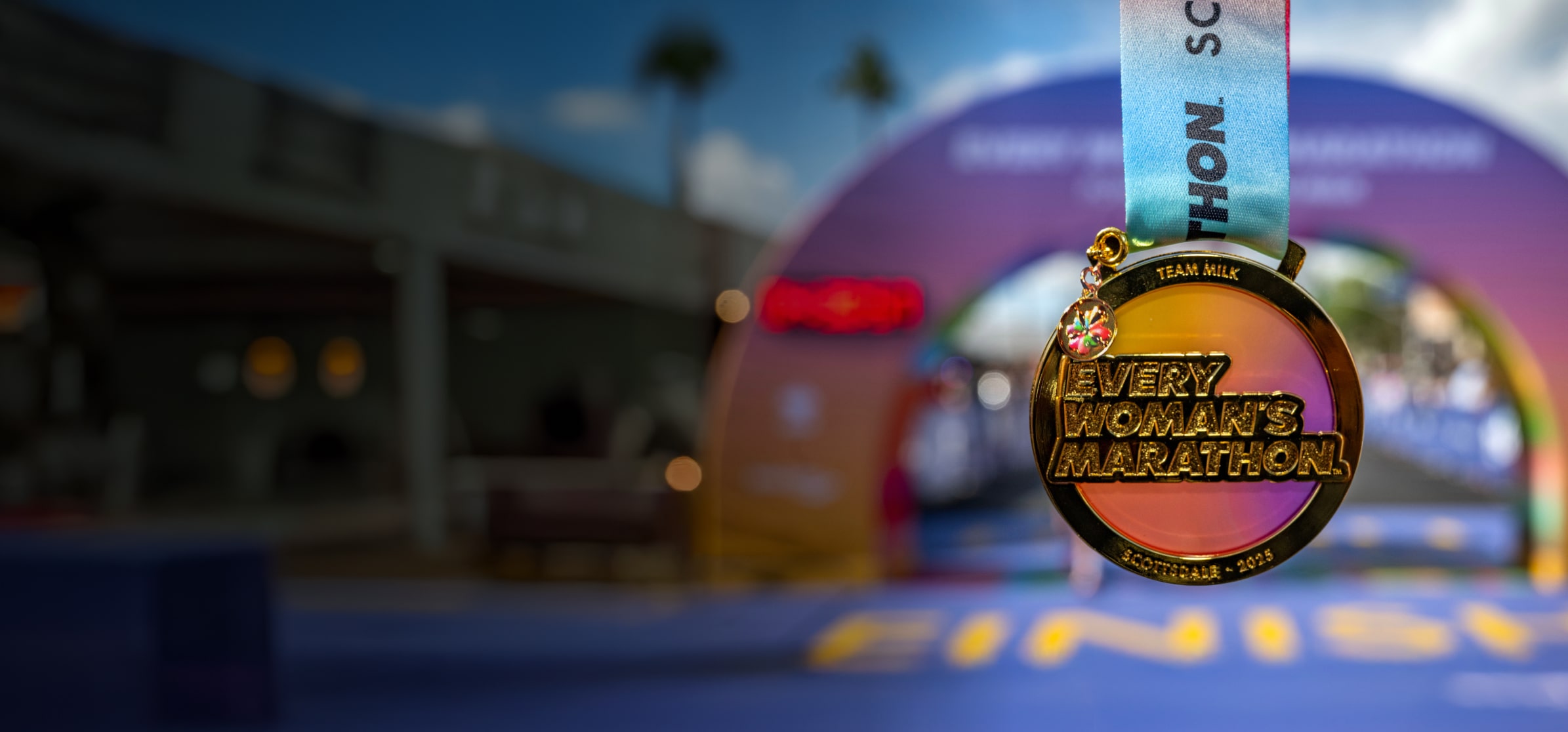 A beautiful gold Every Woman’s Marathon Scottsdale 2025 medal glimmers with accents of a smooth orange and purple gradient in front of a horizon line of the Sonoran Desert.