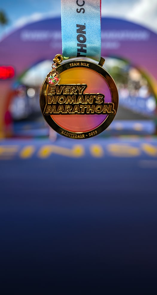 A beautiful gold Every Woman’s Marathon Scottsdale 2025 medal glimmers with accents of a smooth orange and purple gradient in front of a horizon line of the Sonoran Desert.