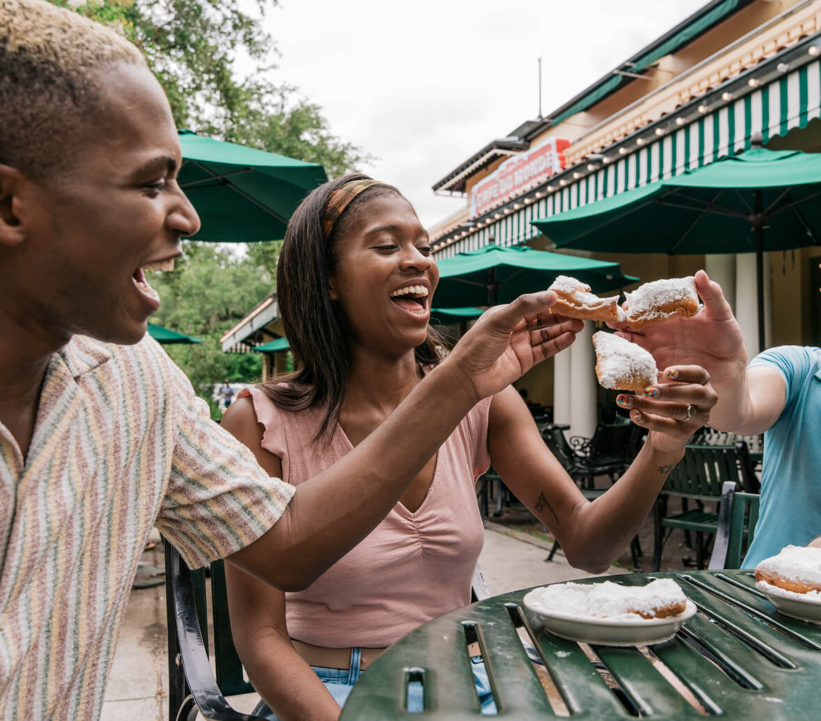 A group of friends cheers with beignets covered in powdered sugar on an outside patio.