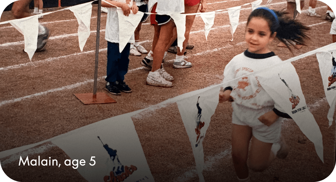 Malain, age 5, runs on a track during a children's race, wearing a white event T-shirt and shorts, with pennant flags and other children in the background. 