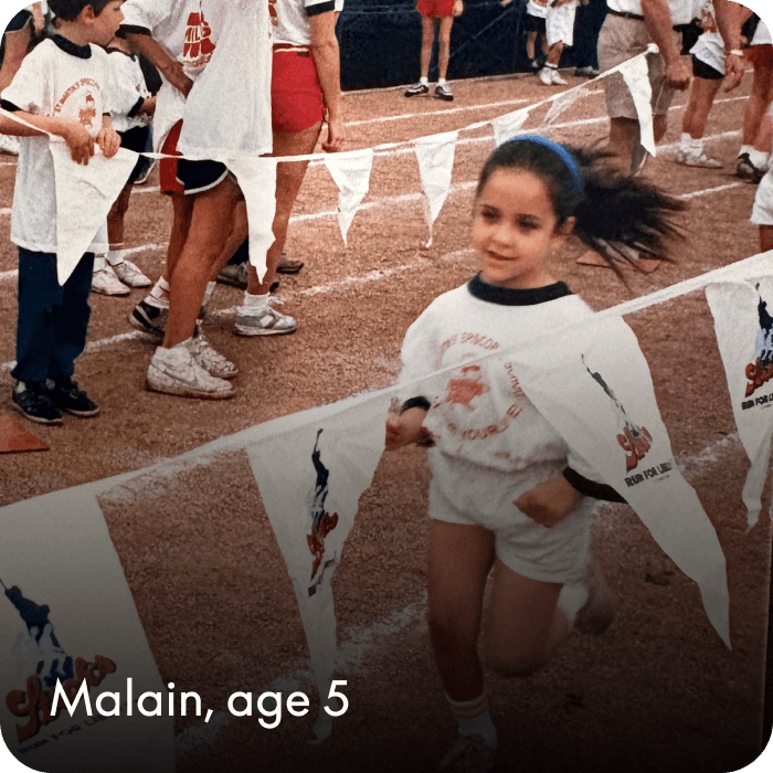 Malain, age 5, runs on a track during a children's race, wearing a white event T-shirt and shorts, with pennant flags and other children in the background. 