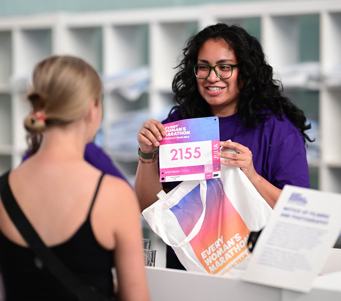 An Every Woman's Marathon volunteer holds up a personalized runner's bib and colorful tote bag during packet pickup.