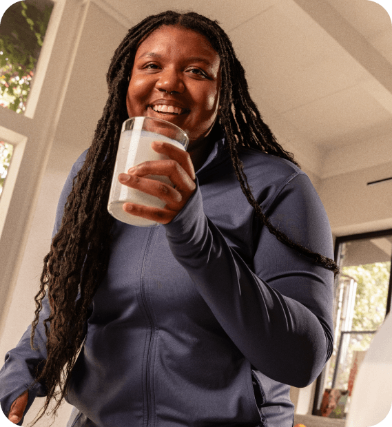 A smiling woman with long locs, wearing a navy athletic jacket and holding a glass of white milk indoors.