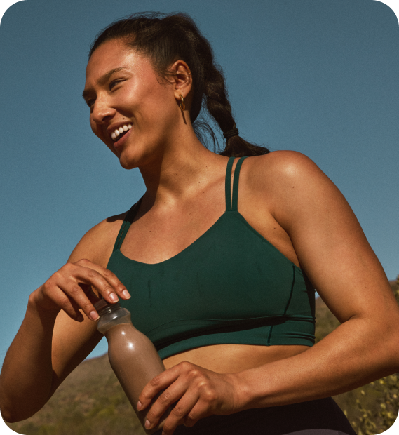 IMG (2) A smiling woman in a green sports bra holds a bottled chocolate milk outdoors beneath a clear blue sky.